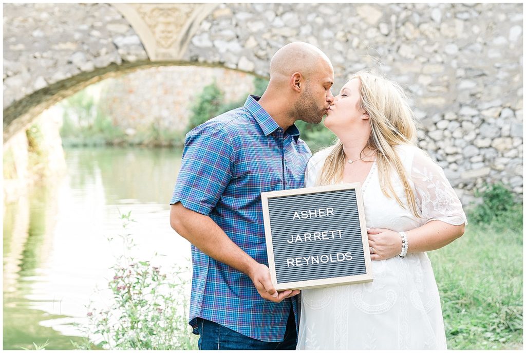pregnant husband and wife kissing at adriatica mckinney name board stone archway river