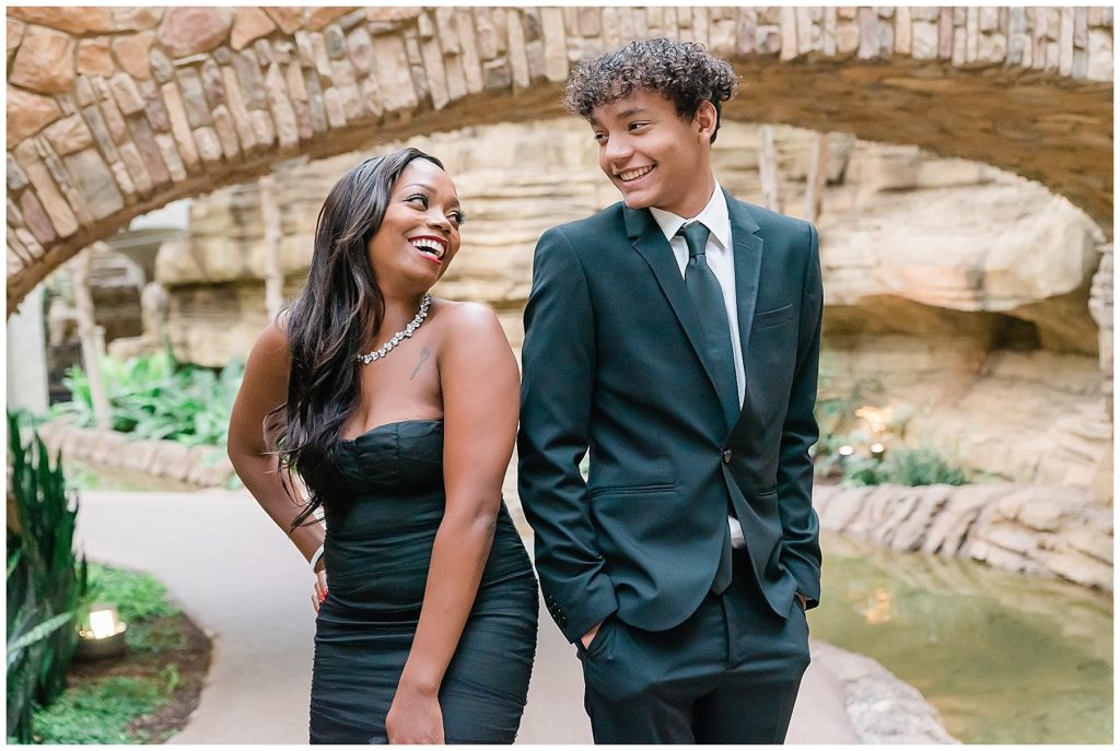 mother in black dress laughing with son in tuxedo on riverwalk