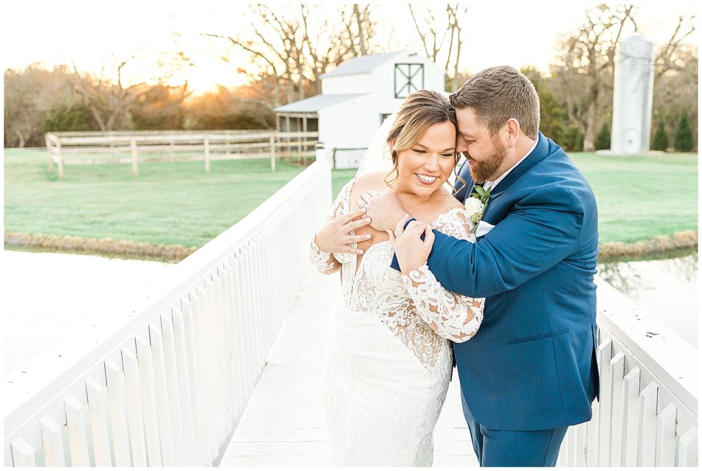 bride in lace gown and groom in navy suit on white bridge in front of white barn
