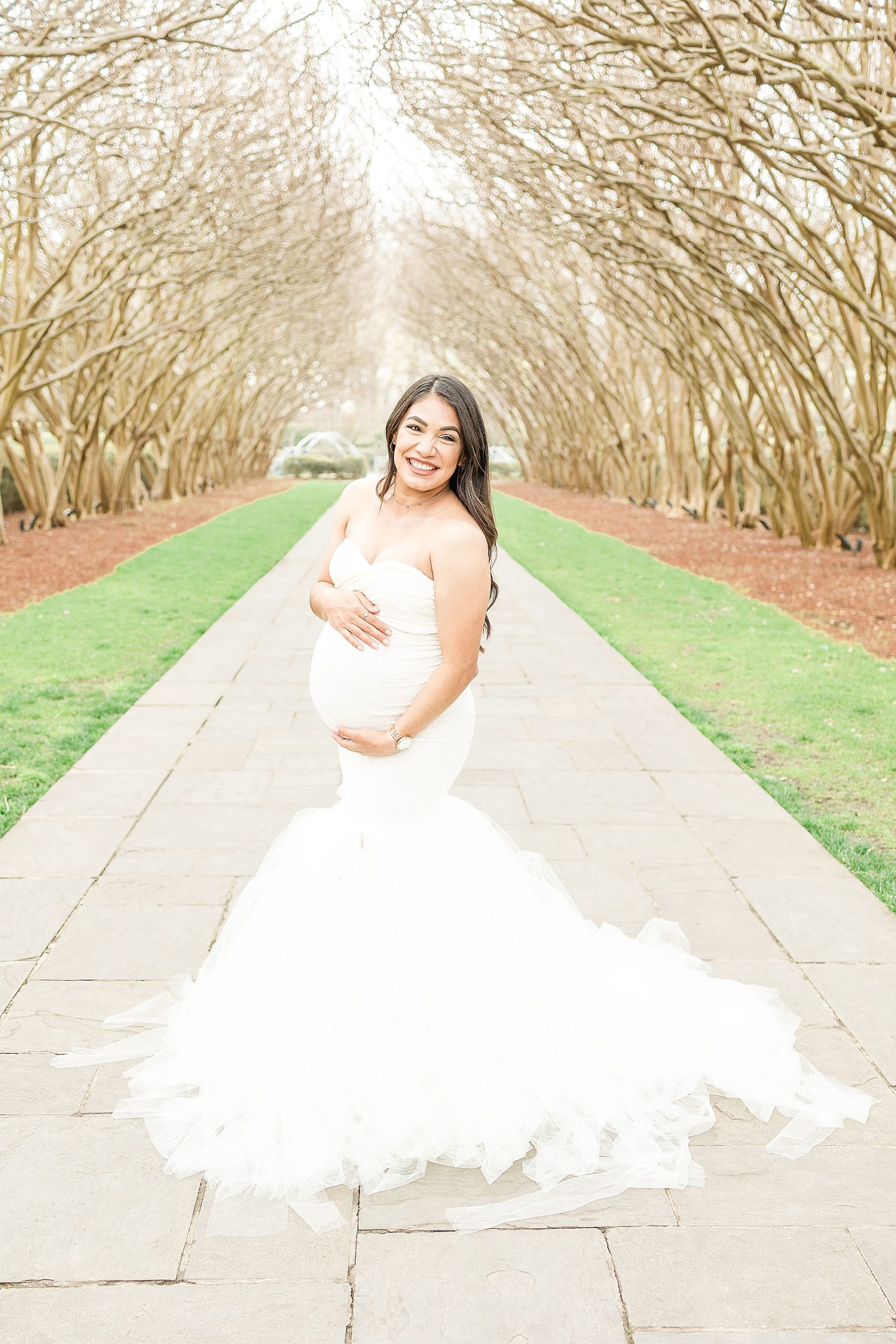 mom in white gown holds belly during Dallas Arboretum maternity session