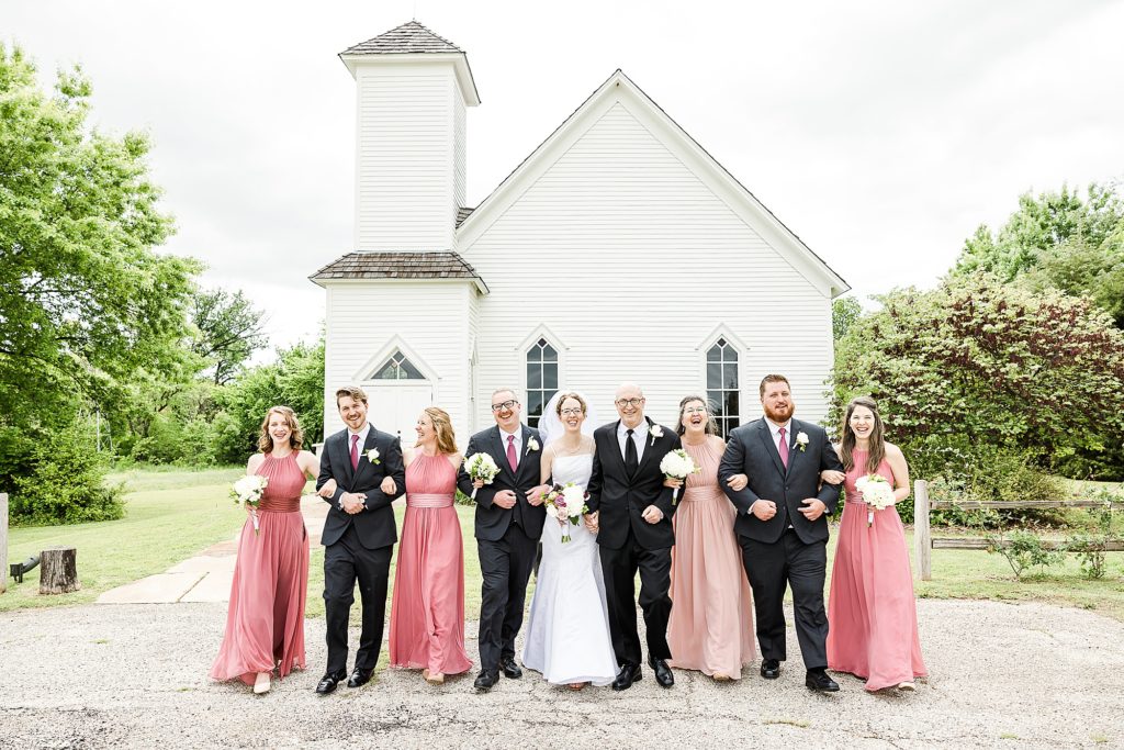 couple walks with wedding party by small white church