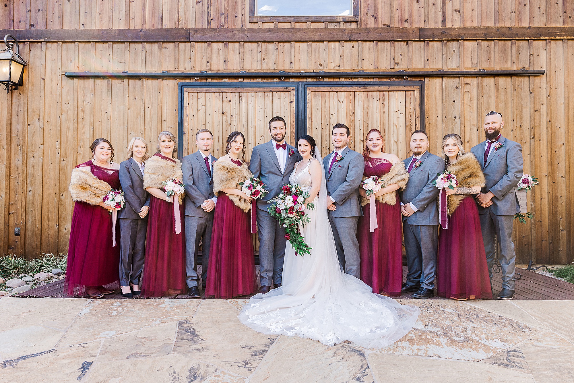 bride and groom pose with wedding party in grey and burgundy outside wooden doors