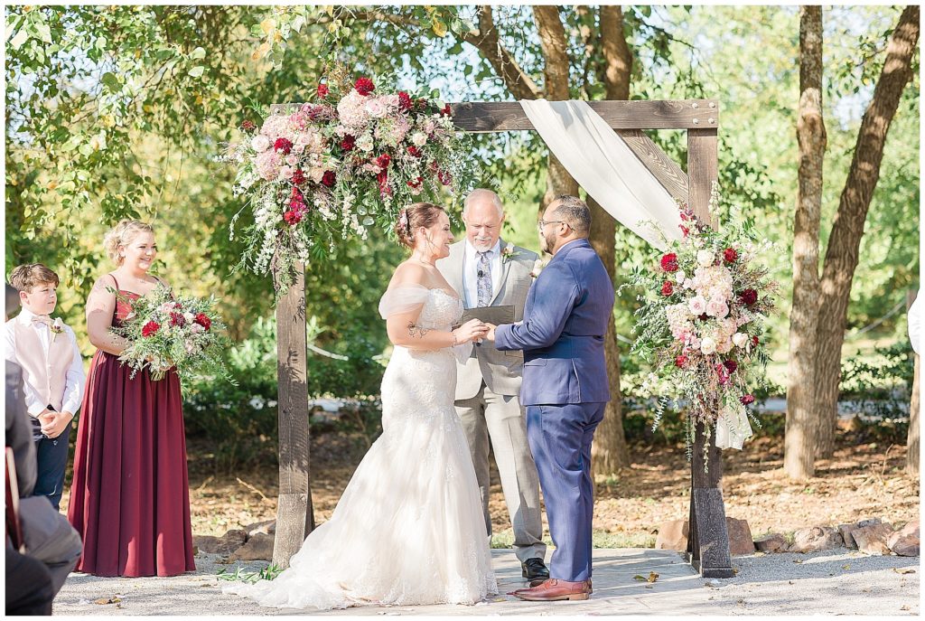 bride and groom exchanging rings under rustic arch with dahlia and rose florals