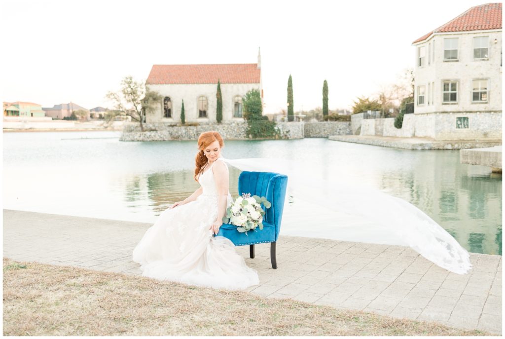 bride with cathedral length cape veil on blue velvet chair by lake in front of stone church