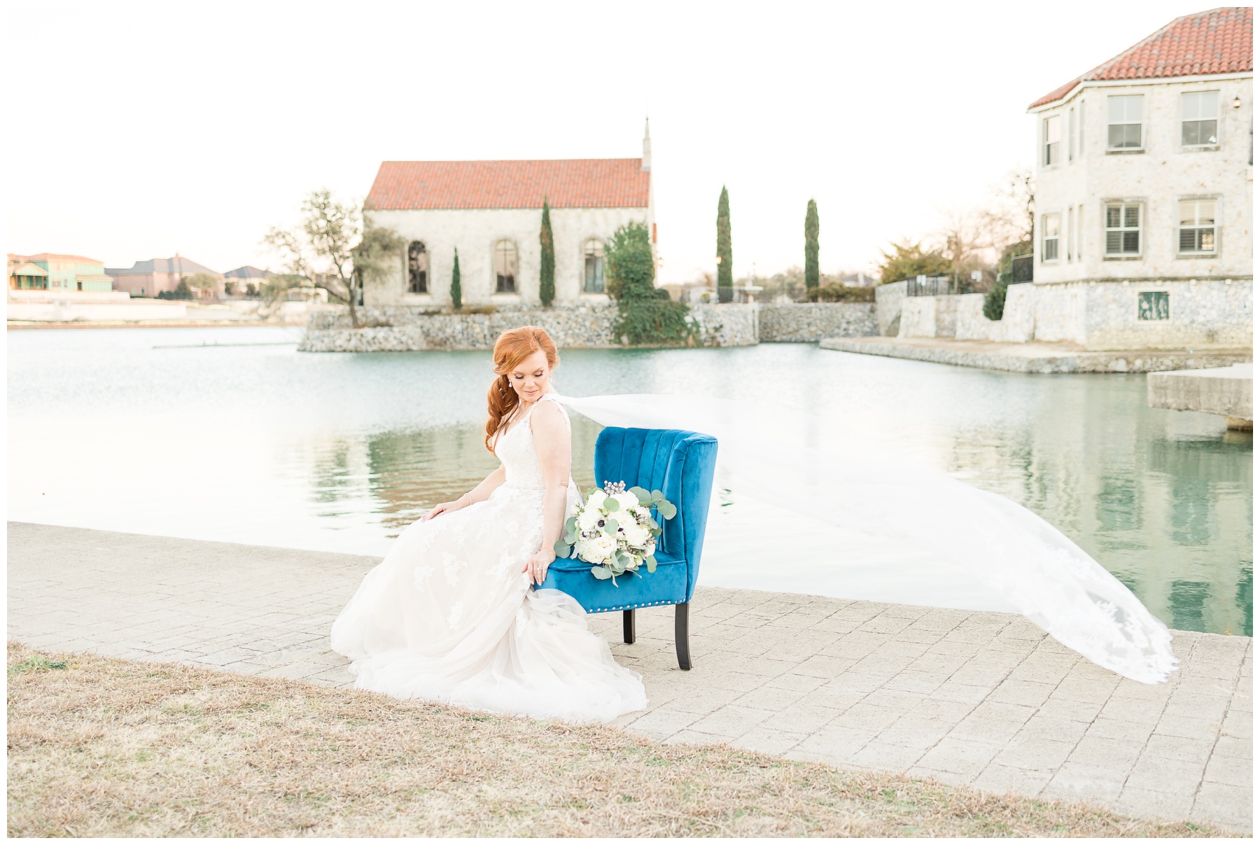 bride with shoulder cape veil sitting on blue velvet chair in front of lake and white stone church