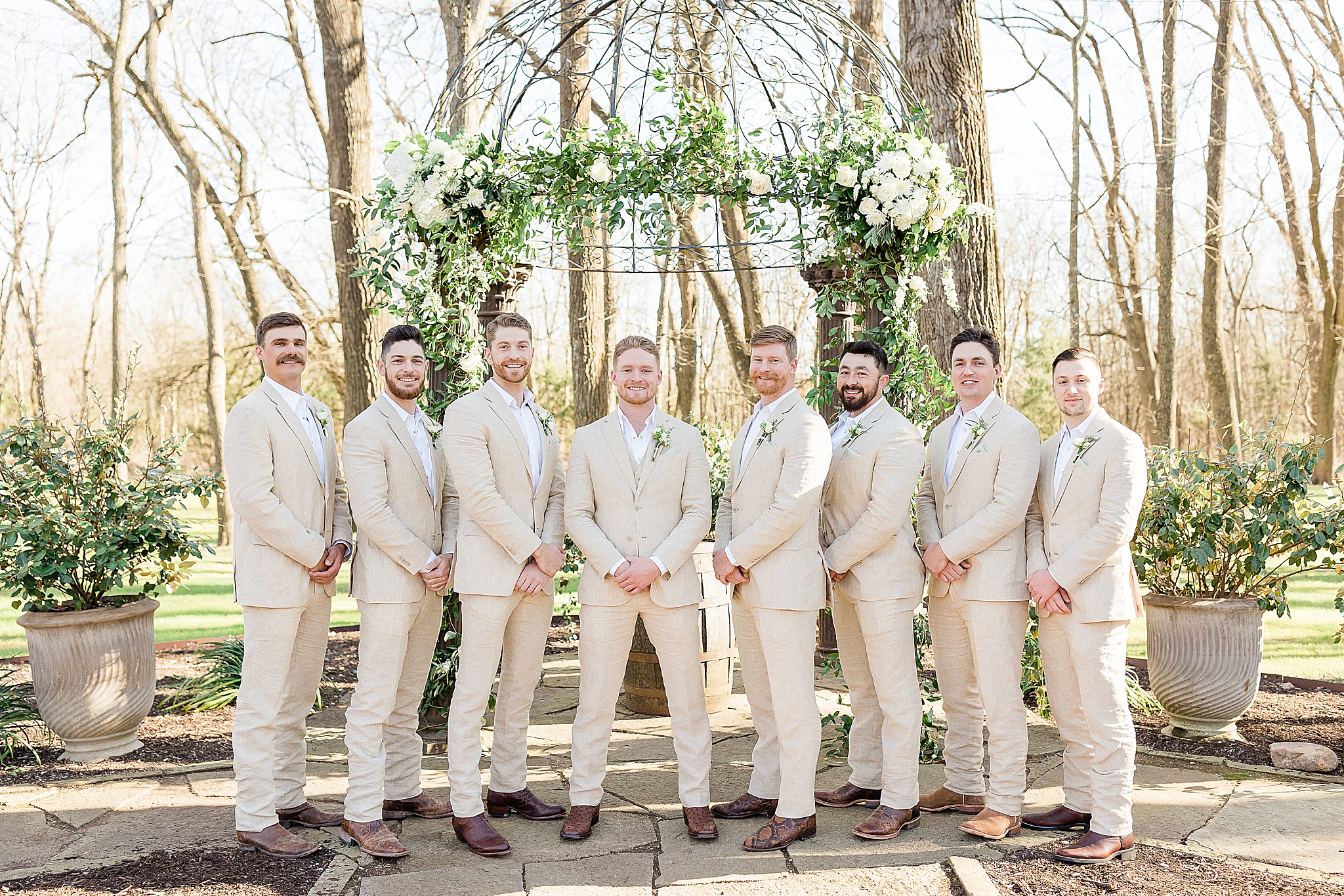 groom and groomsmen in tan suits pose by floral arbor 