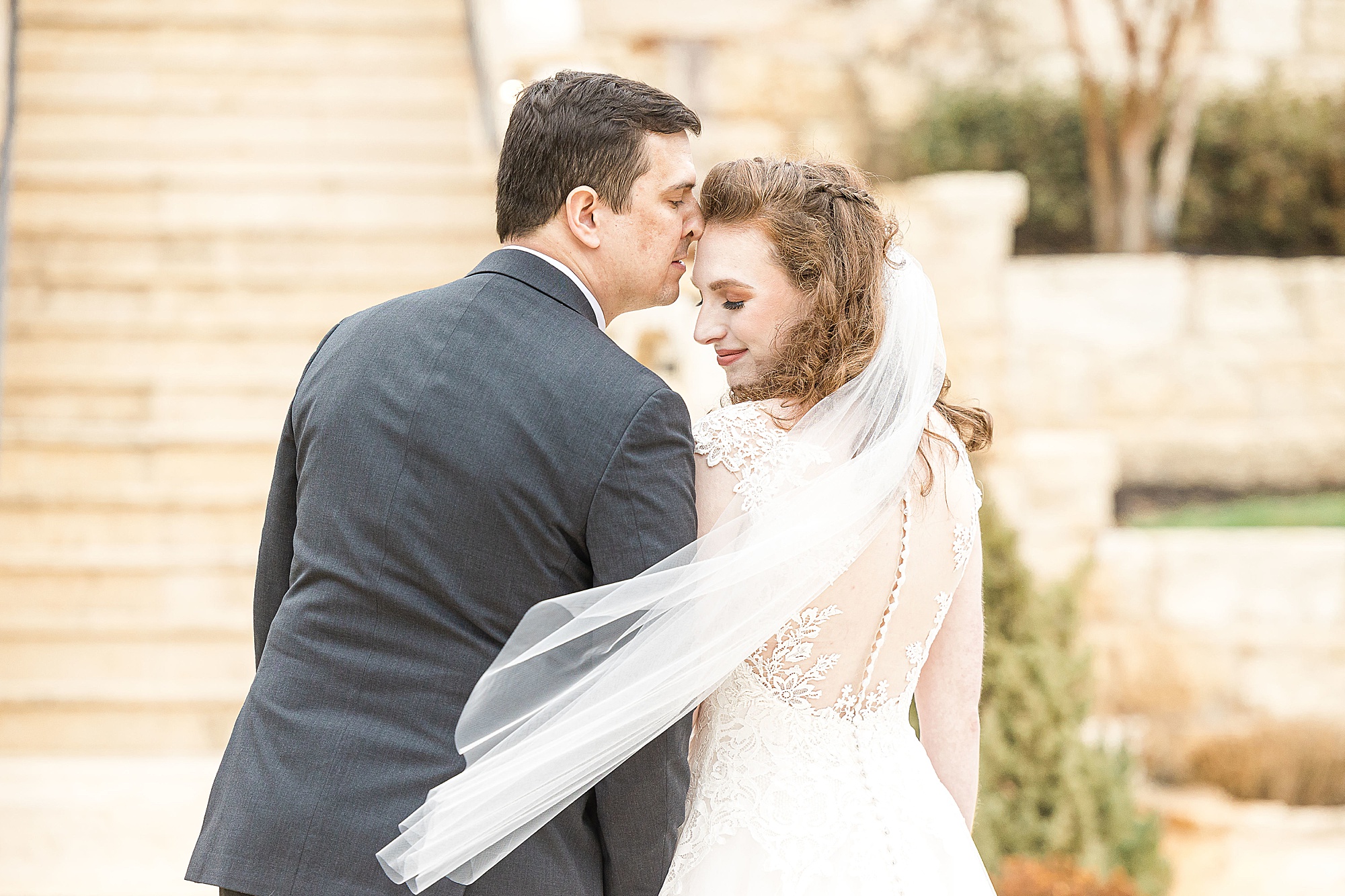 groom kisses bride's forehead while veil floats behind her