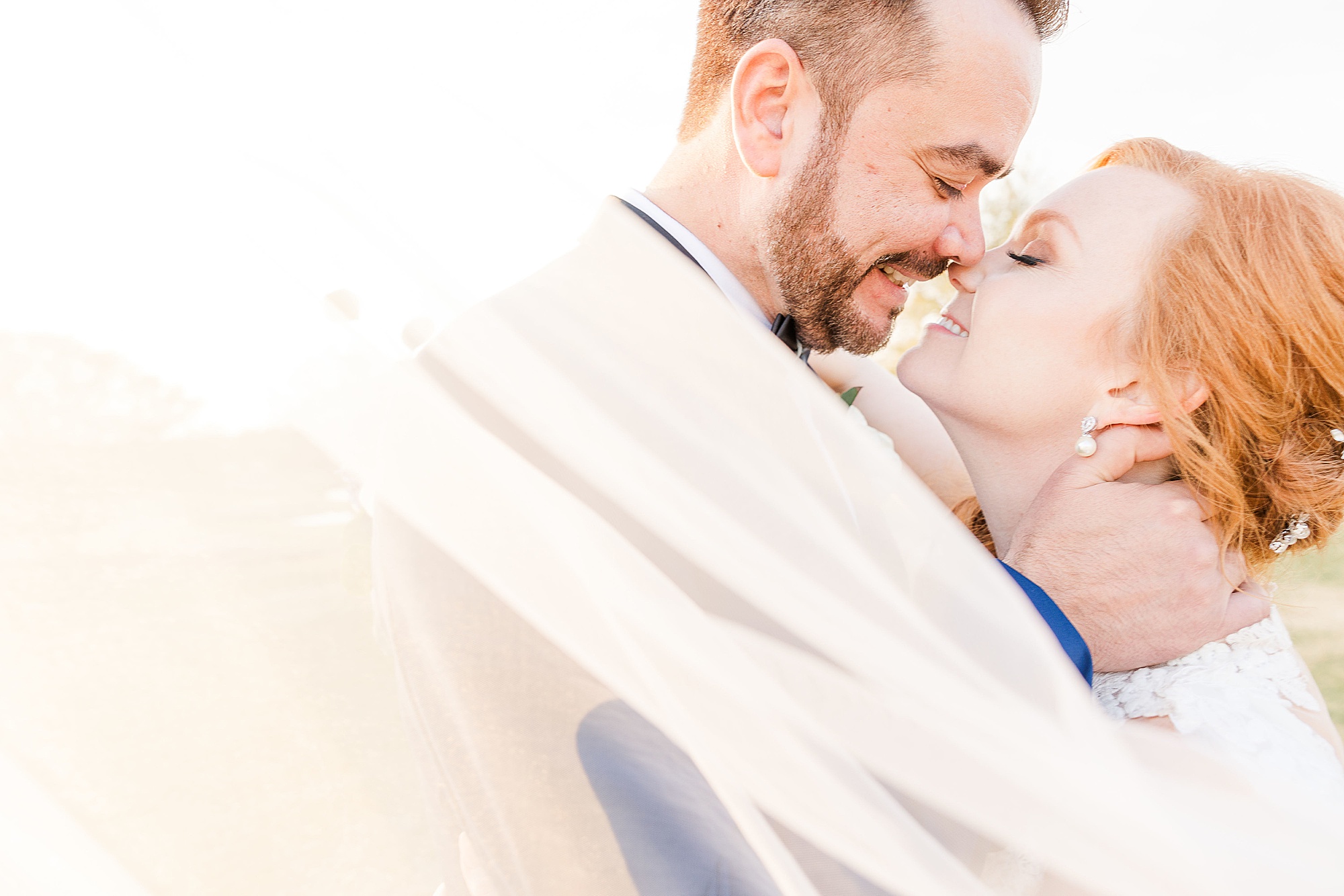 bride and groom stand touching noses with veil around them